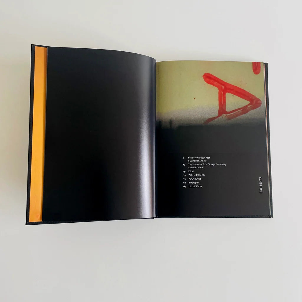 An open hardcover book or album on a light gray background, showing a mostly black cover with a bright orange vertical stripe on the left page and a printed interior spread with a red abstract graphic and track list on the right page.