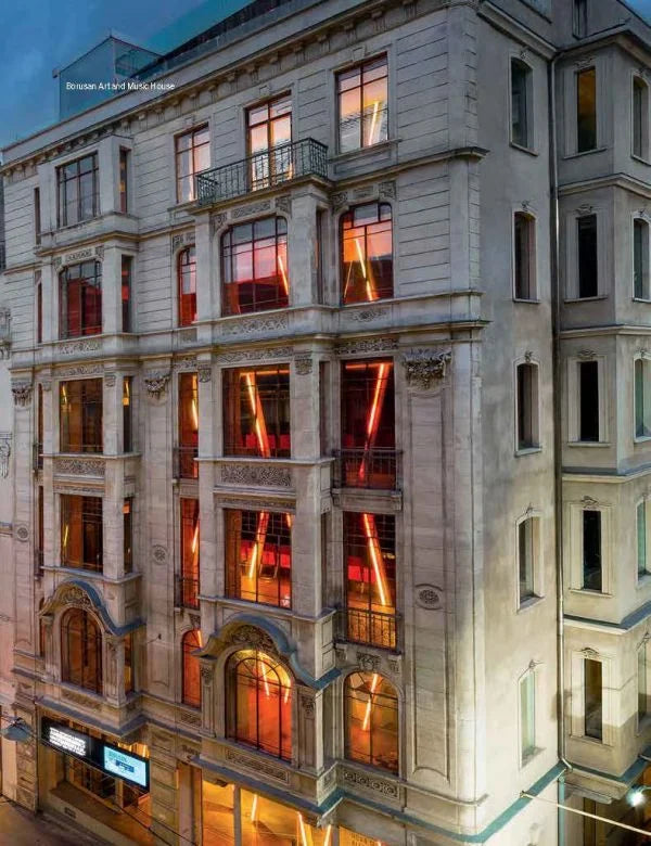 An ornate multi-story stone building facade at dusk, with warm orange-red lights glowing through many windows and a pale evening sky above.