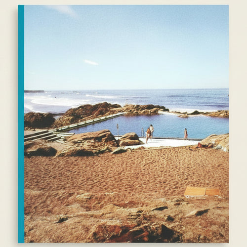 Natural rock pool with people around, surrounded by sandy beach and ocean.