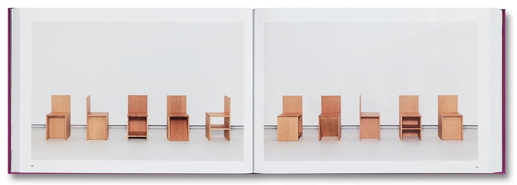 An open book or art print showing a minimalist interior scene with several small wooden chairs arranged across two white pages. The chairs are light tan and medium brown wood tones, set against a clean white background.