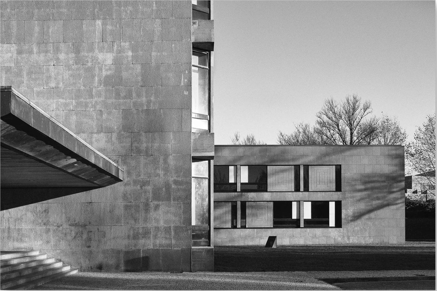 Black and white architectural photograph of a modernist concrete building with geometric forms, strong shadows, and bare trees in the background.