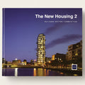 A book cover titled "The New Housing 2: Building Better Communities" featuring a twilight photograph of a modern illuminated apartment building beside water, with the RIA logo in the lower right.