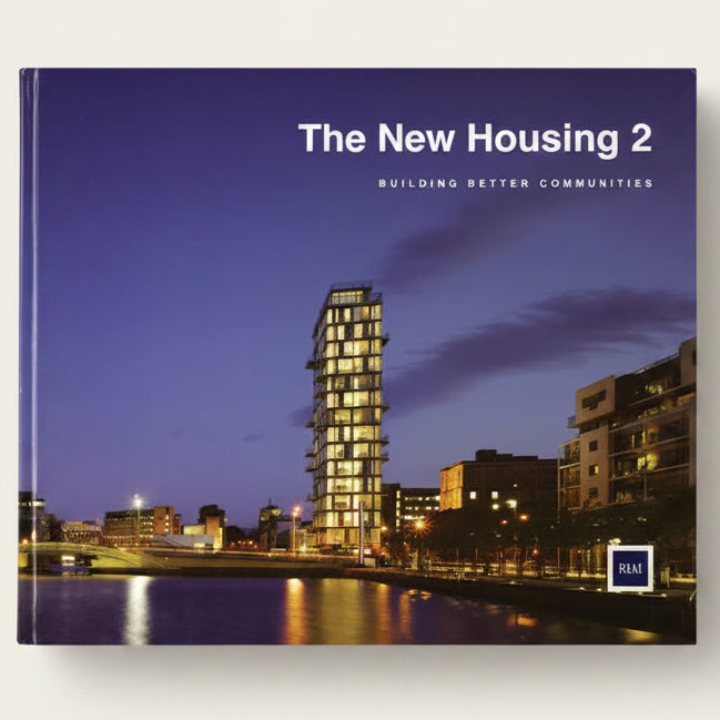 A book cover titled "The New Housing 2: Building Better Communities" featuring a twilight photograph of a modern illuminated apartment building beside water, with the RIA logo in the lower right.