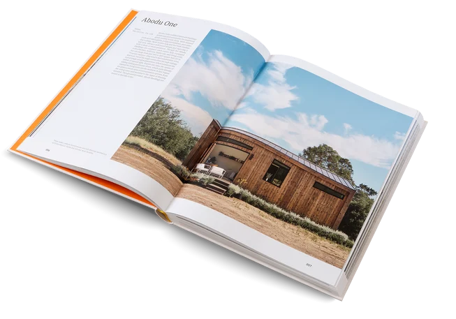 An open hardcover book showing a large architectural photograph of a wooden cabin on the right page and text on the left page, photographed on a clean white background.