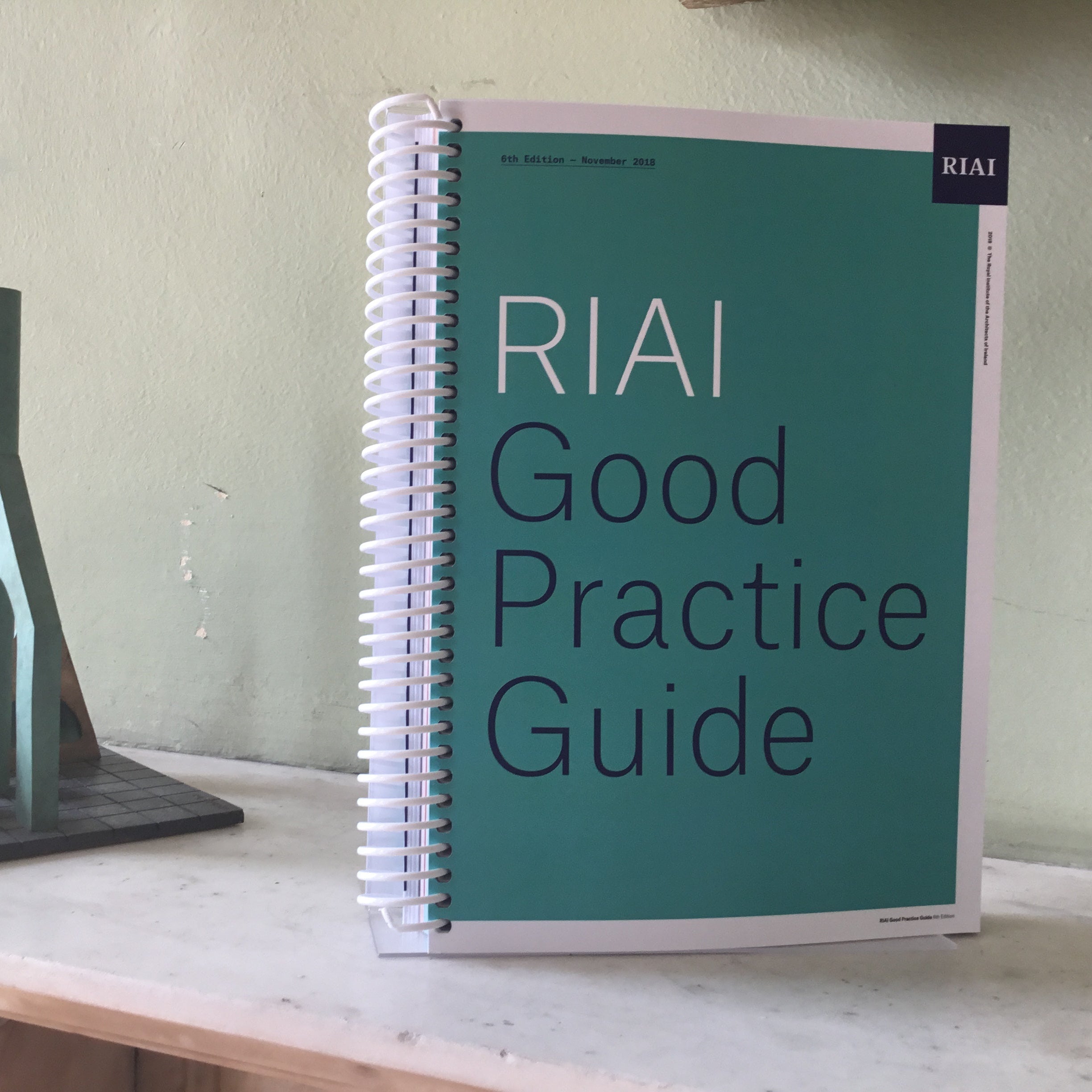 Spiral-bound book titled 'RIAI Good Practice Guide' on a table with a model of a building.