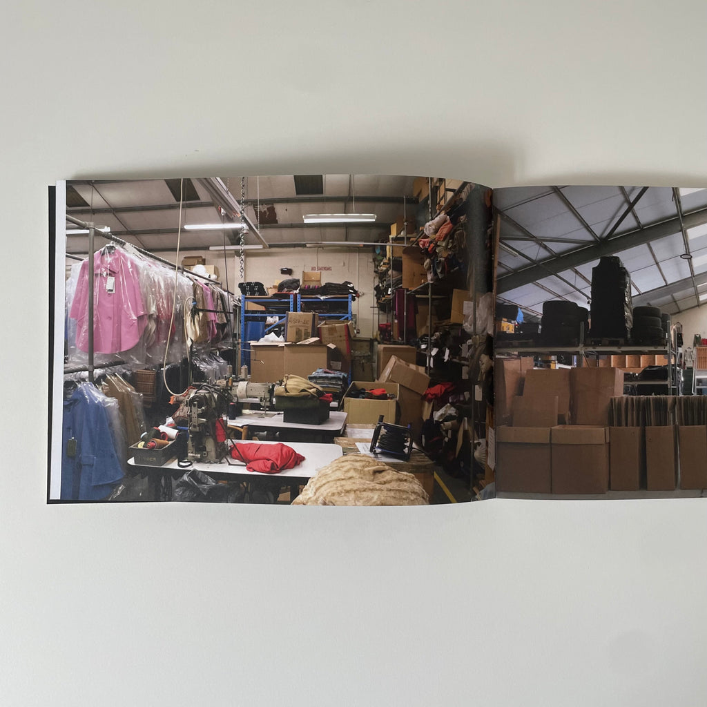 An open book or magazine spread photographed against a plain white wall. The pages show a warehouse or retail interior with clothing racks, shelves, boxes, and work tables.