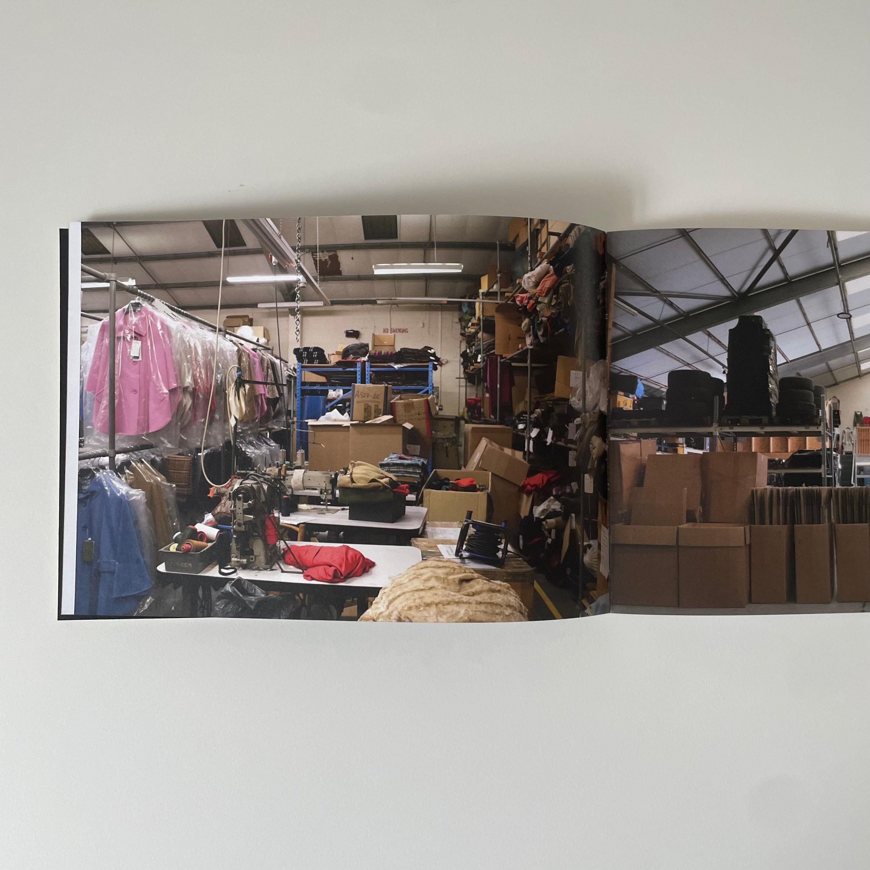 An open book or magazine spread photographed against a plain white wall. The pages show a warehouse or retail interior with clothing racks, shelves, boxes, and work tables.