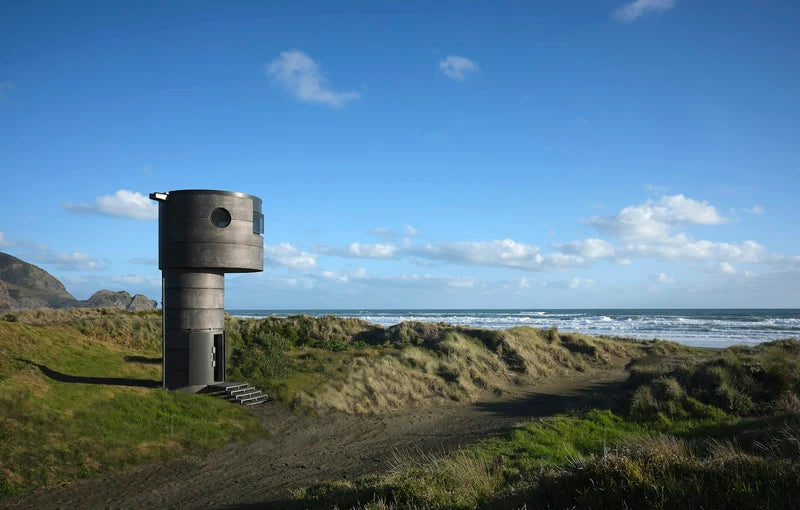 A cylindrical metal structure, possibly a small tower or container, standing on a grassy dune next to the ocean under a bright blue sky with scattered clouds.